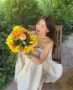 a woman sitting on a bench holding a bouquet of yellow and orange flowers in her hands