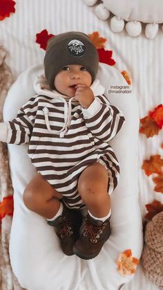 a baby in a striped shirt and hat is sitting on a blanket with autumn leaves