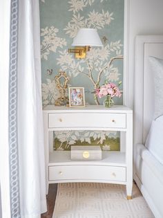 a white dresser sitting next to a bed in a room with floral wallpaper on the walls