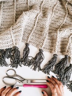 a woman's hands holding scissors next to a knitted blanket with tassels