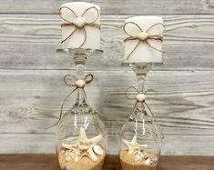 two glass vases with sand and shells in them on a wooden table next to each other