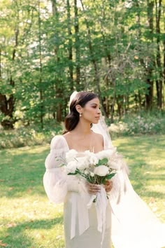 a woman in a wedding dress holding a bouquet and looking off into the distance with trees in the background