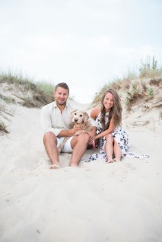 a man and woman are sitting on the sand with their dog in front of them