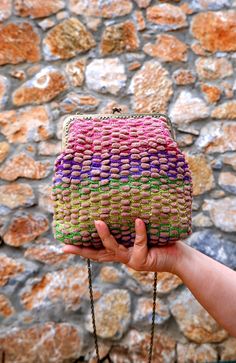 a hand holding a colorful woven purse in front of a stone wall