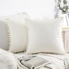 two white pillows sitting on top of a couch next to a pillow with pom - poms