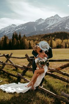 a bride and groom kissing in front of a wooden fence with mountains in the background