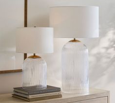 two clear glass lamps sitting on top of a wooden dresser next to a white lamp shade