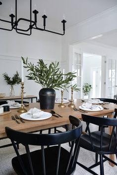 a dining room table with black chairs and white plates on top of it, in front of a potted plant