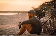 a man sitting on the beach next to his bike with a beer in front of him