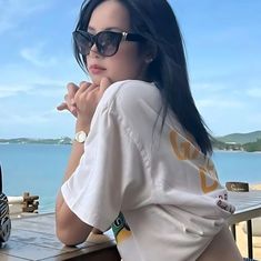 a woman wearing sunglasses sitting on a bench next to the ocean with her hand under her chin