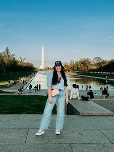 a woman standing in front of the washington monument wearing jeans and a baseball cap with her hand on her hip