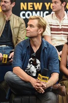 a man sitting in front of a microphone with other people behind him at a comic convention