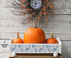 three pumpkins are sitting in a white tray on a wooden table next to a dried twig wreath