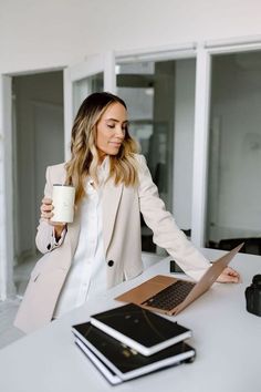 a woman sitting at a desk holding a coffee cup and looking at her laptop computer