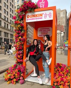 two women standing in a phone booth with flowers on the ground and buildings behind them