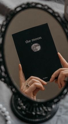 a person holding up a black book in front of a mirror with the moon on it