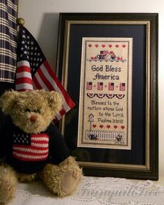 a teddy bear sitting next to an american flag and a framed cross - stitch sample