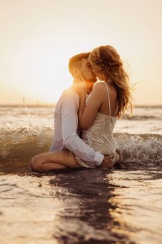 two people sitting on the beach kissing each other and having fun in the water at sunset