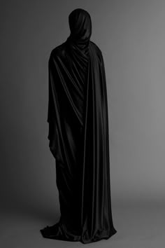 a woman wearing a black cloak and standing in front of a gray background with her back turned to the camera