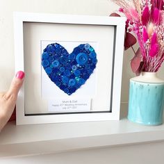 a blue heart made out of buttons in a white frame next to a vase with pink flowers