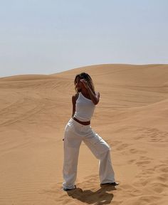 a woman is standing in the sand with her hands behind her head and wearing white pants