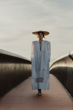 a woman walking across a bridge wearing a white dress and a large hat on her head