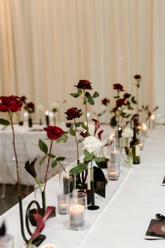 several vases with roses and candles are lined up on a long white table cloth