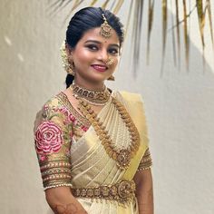 a woman in a white and gold sari with jewelry on her neck, standing next to a palm tree