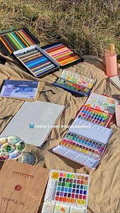 an assortment of art supplies laid out on a blanket in the grass, including watercolors and pencils