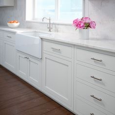 a kitchen with white cabinets and wooden floors, pink flowers in a vase on the window sill