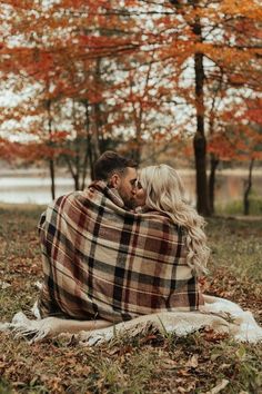 a man and woman wrapped up in a blanket sitting on the ground with trees behind them