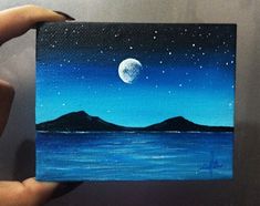 a person holding up a small painting with the moon in the sky above water and mountains