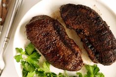 two steaks on a white plate with parsley and silverware next to it