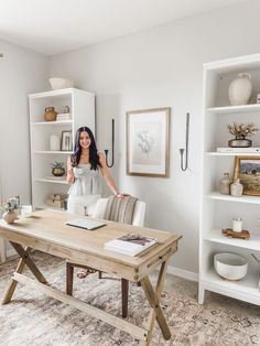 a woman standing in front of a desk with books on it and shelves behind her