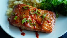 a white plate topped with salmon and broccoli covered in sauce next to rice