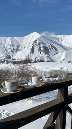 two cups of coffee sit on a balcony overlooking the snow - covered mountains and valleys