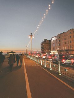 people are walking on the sidewalk next to some parked cars and street lights at night