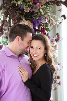 a man and woman standing next to each other in front of a flower covered wall