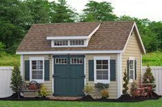 a small shed sitting in the grass next to a white fence and some bushes with potted plants