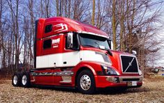 a red and white semi truck parked in front of some trees with no leaves on the ground