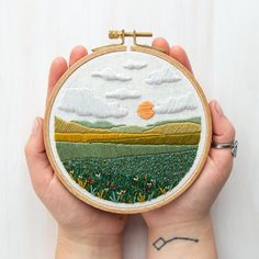 a person holding up a small embroidery hoop with an image of a field in the background