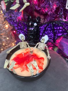 two skeletons sitting in a black bowl with red liquid on the surface and purple decorations behind them