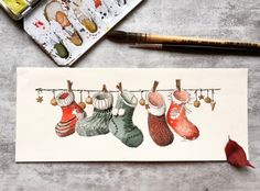 a watercolor painting of christmas stockings hanging on a clothes line with paintbrushes next to it