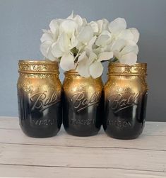 three black mason jars with gold lids and white flowers in them on a wooden table