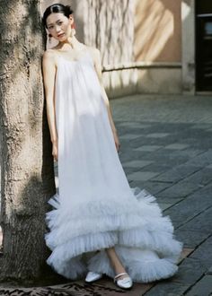 a woman standing next to a tree wearing a white dress with ruffles on it