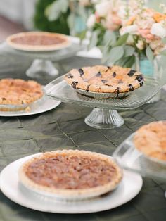 several pies on plates sitting on a table with flowers in vases behind them