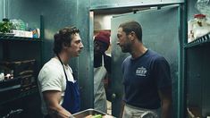 two men standing in a kitchen talking to each other while another man looks at him