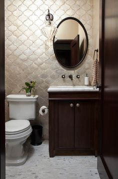 a white toilet sitting next to a sink in a bathroom under a round mirror above a wooden cabinet