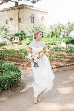 a woman in a white dress holding a bouquet and walking down a path with an old building in the background