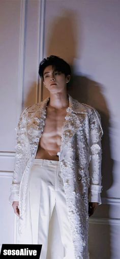 a shirtless man leaning against a wall wearing white pants and a jacket with flowers on it